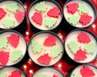 Könnte beinhalten: Eine Sammlung kleiner, runder Kerzen in schwarzen Dosen. Jede Kerze hat eine cremeweiße Wachsbasis, verziert mit roten und grünen Weihnachtsbaum-förmigen Wachsverzierungen. Die Kerzen sind auf einer roten Oberfläche angeordnet.