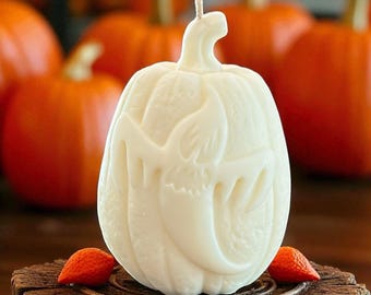 Puede incluir: Una vela con forma de calabaza de color crema con una cara de Jack-o'-lantern tallada. Dos pequeñas decoraciones naranjas en forma de lágrima se encuentran en la base de la vela. El fondo presenta varias calabazas naranjas.