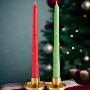Puede incluir: Dos velas cónicas, una roja y otra verde, cada una con un diseño de árbol de Navidad, se colocan en soportes dorados. Las velas están frente a un árbol de Navidad borroso con adornos y luces.