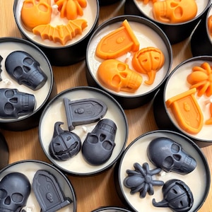 Pode incluir: Uma coleção de velas com tema de Halloween em latas de metal preto. Cada vela apresenta uma figura de cera diferente, incluindo caveiras, abóboras, morcegos e lápides. As figuras de cera são laranja e pretas, com uma base de vela branca cremosa.