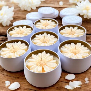 May include: A collection of daisy-shaped candles in light blue tins with gold rims. The candles are a creamy yellow color and have a single wick. White lids are visible in the background, along with white flower petals.