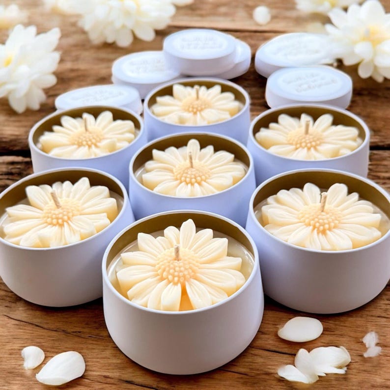 May include: A collection of daisy-shaped candles in light blue tins with gold rims. The candles are a creamy yellow color and have a single wick. White lids are visible in the background, along with white flower petals.