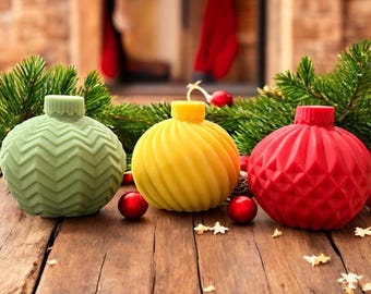 Puede incluir: Tres adornos navideños decorativos en verde, amarillo y rojo, cada uno con un diseño texturizado único. Los adornos están dispuestos sobre una superficie de madera con agujas de pino y bayas rojas, creando una escena festiva navideña.
