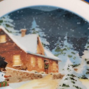 Puede incluir: Un plato de cerámica blanco con borde dorado presenta una escena de invierno con un muñeco de nieve, una casa y árboles. El texto "All roads lead home at Christmas" está impreso en el plato.
