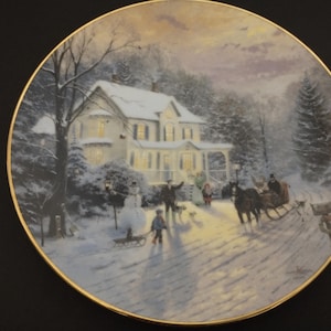Puede incluir: Un plato de porcelana blanca con borde dorado presenta una escena de invierno con una casa cubierta de nieve, un trineo tirado por un caballo y figuras caminando en la nieve. La escena está iluminada por una luz cálida que proviene de las ventanas de la casa.