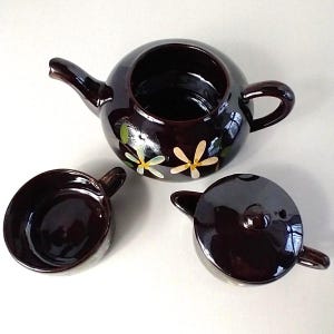 May include: A dark brown ceramic teapot set with a matching lid and cup. The teapot features a round body, a spout, and a handle. The teapot and cup are decorated with hand-painted yellow and white floral designs.