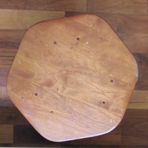 May include: A top-down view of a pentagonal wooden stool seat featuring a light brown natural wood grain finish. The surface displays five dark circular screw or nail heads arranged in a pentagonal pattern, with visible wood texture and grain.