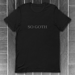 May include: Black t-shirt featuring the text "SO GOTH" in grey, gothic lettering. The tee has a crew neck and short sleeves. The background shows a dark wood grain pattern.
