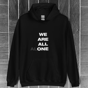 Puede incluir: Sudadera con capucha negra con el texto "WE ARE ALL ALONE" en blanco y gris. La sudadera tiene un bolsillo delantero y una capucha. La marca "Gildan" es visible en la etiqueta.