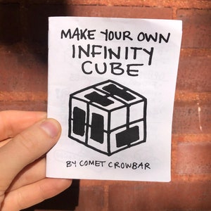 May include: A white booklet with black text and a black and white illustration of a cube. The text reads "Make Your Own Infinity Cube" and "By Comet Crowbar".