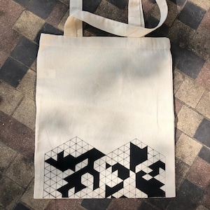 May include: A white canvas tote bag with black geometric design printed on the bottom. The design is made up of triangles and squares.