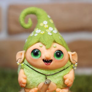 May include: A green felt gnome with a curly green felt hat, green eyes, and a happy expression. The gnome is wearing a beaded necklace and has a small wooden stick in its hand.