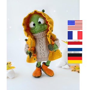 May include: A crocheted frog wearing a yellow raincoat, a beige sweater, and orange boots. The frog is holding a small orange leaf in its hand.