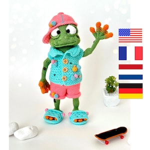 May include: A crocheted frog wearing a pink hat, blue shirt, pink shorts, and blue slippers. The frog is holding its right hand up in a wave. The frog is standing on a white surface with a small skateboard in the foreground.