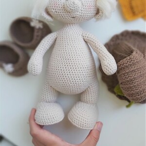 May include: A crocheted doll with white yarn hair, brown eyes, and a beige body. The doll is standing on a white surface.