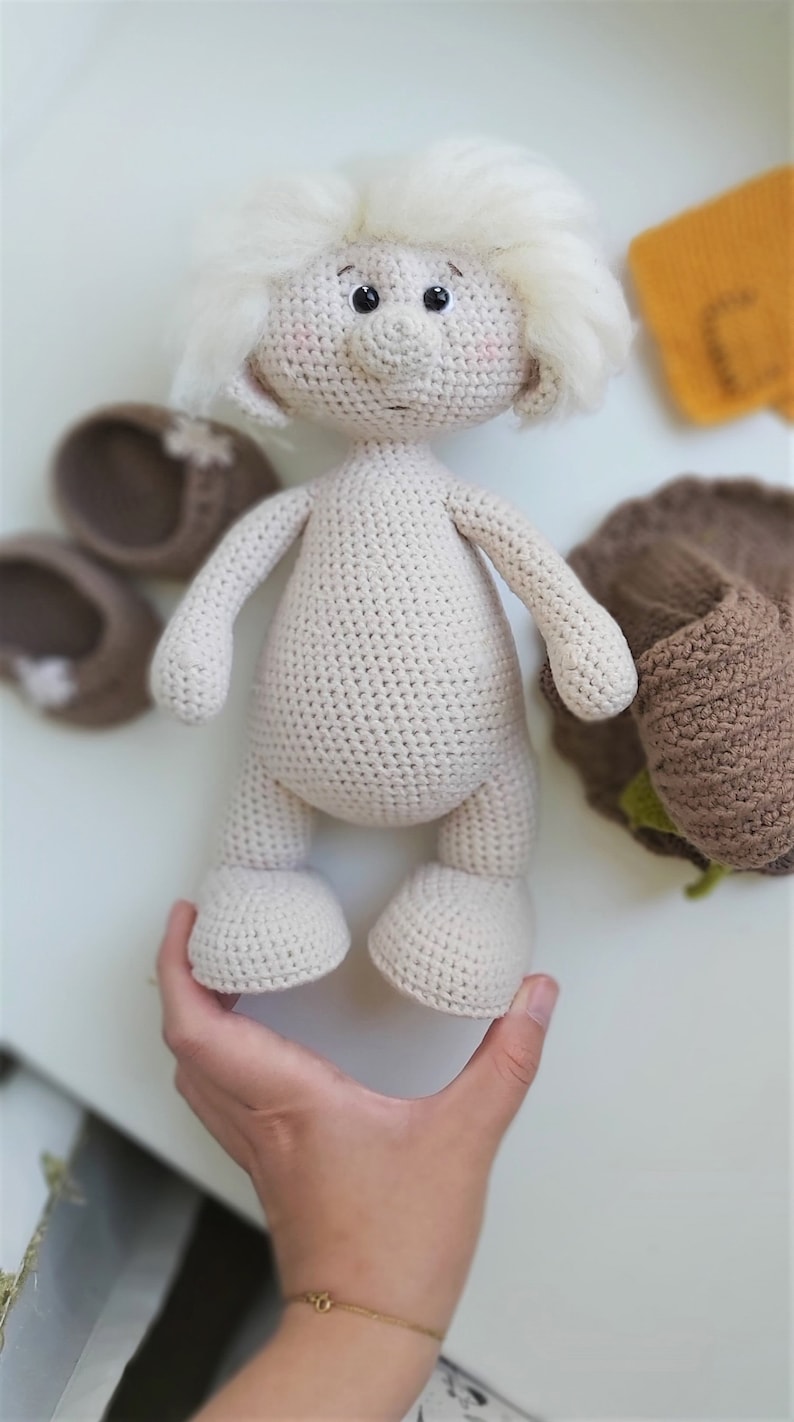 May include: A crocheted doll with white yarn hair, brown eyes, and a beige body. The doll is standing on a white surface.