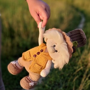 May include: A crocheted gnome doll wearing a yellow vest, brown hat, and brown shoes. The gnome has a white beard and brown eyes.