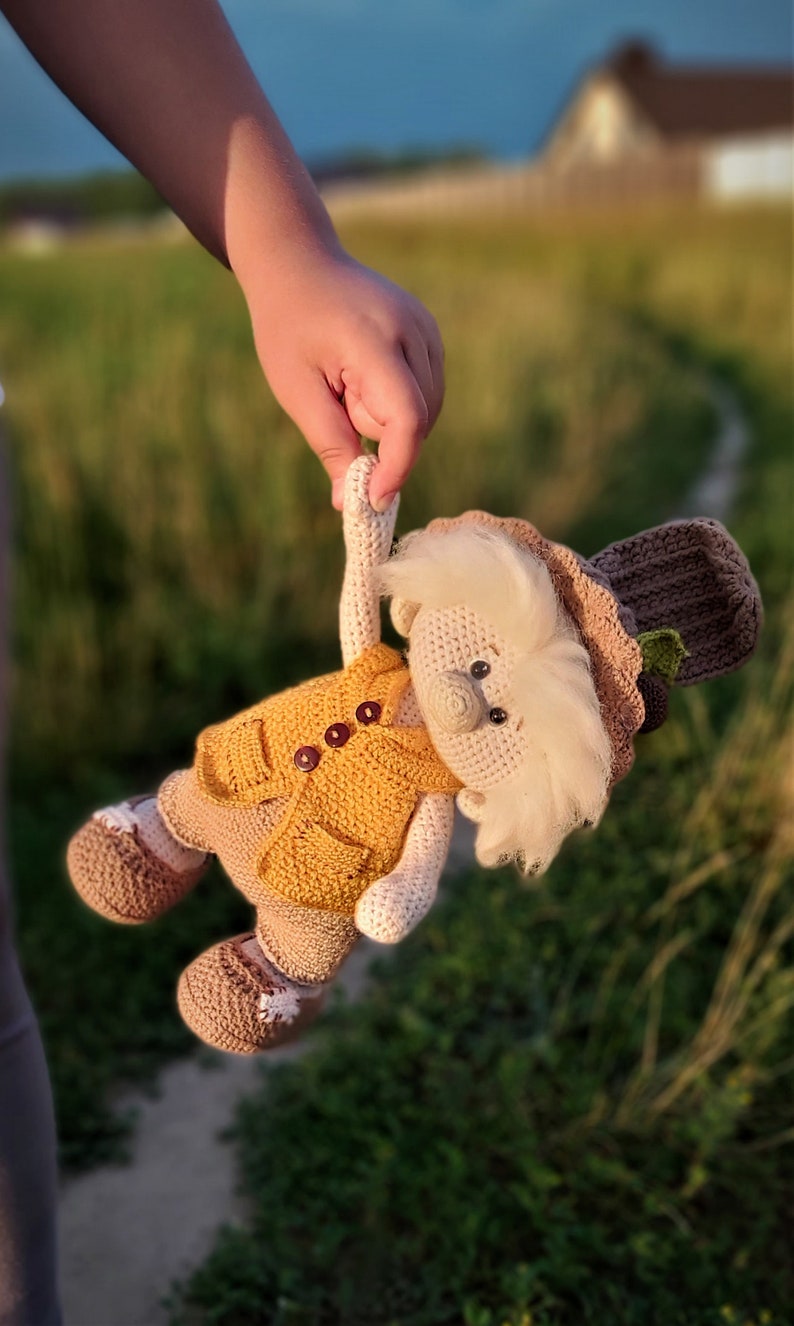 May include: A crocheted gnome doll wearing a yellow vest, brown hat, and brown shoes. The gnome has a white beard and brown eyes.