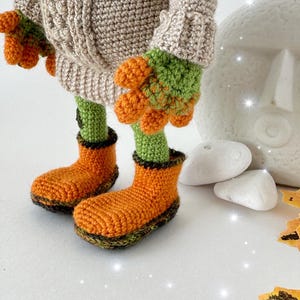 May include: A crocheted toy wearing a beige sweater with green and orange accents. The toy is wearing orange boots with green trim. The toy is standing on a white surface with white stones and orange leaves.