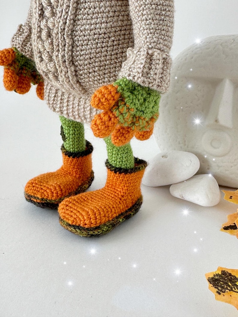 May include: A crocheted toy wearing a beige sweater with green and orange accents. The toy is wearing orange boots with green trim. The toy is standing on a white surface with white stones and orange leaves.