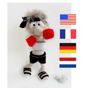 Könnte beinhalten: Eine gehäkelte Boxerpuppe mit roten Boxhandschuhen, schwarzen Shorts und weißen Schuhen. Die Puppe hat ein schwarzes Stirnband und braunes Haar. Die Puppe steht auf einer weißen Oberfläche mit drei weißen Bällen im Hintergrund. Im Hintergrund sind vier Flaggen zu sehen: die amerikanische Flagge, die französische Flagge, die deutsche Flagge und die niederländische Flagge.