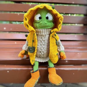 May include: A green crocheted frog wearing a yellow raincoat, a beige sweater, and orange rain boots. The frog is sitting on a wooden bench.