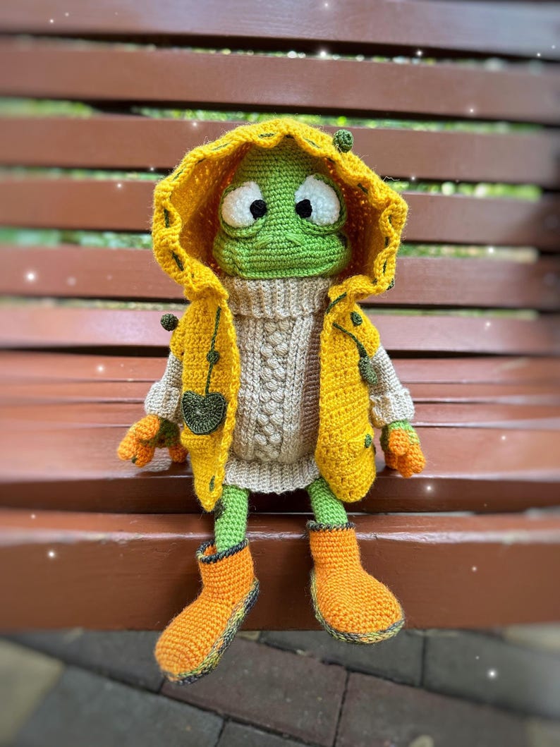 May include: A green crocheted frog wearing a yellow raincoat, a beige sweater, and orange rain boots. The frog is sitting on a wooden bench.