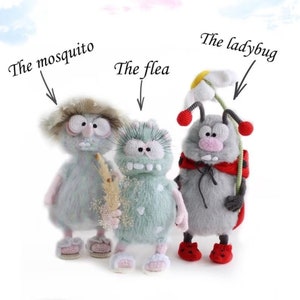 Puede incluir: Tres juguetes de peluche de crochet, un mosquito, una pulga y una mariquita. El mosquito lleva un sombrero y sostiene un palo. La pulga lleva un vestido de lunares verdes y blancos. La mariquita lleva un vestido rojo y negro y una flor blanca en la cabeza. Los juguetes están todos de pie sobre un fondo blanco.