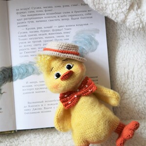 May include: A crocheted yellow chick toy with orange feet, a red bow tie, and a brown and orange hat. The chick is sitting on a white fluffy surface with a book in the background.