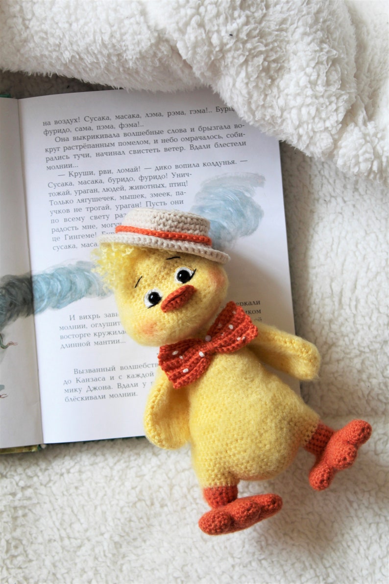 May include: A crocheted yellow chick toy with orange feet, a red bow tie, and a brown and orange hat. The chick is sitting on a white fluffy surface with a book in the background.