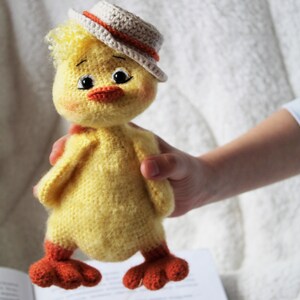 May include: A crocheted yellow chick toy wearing a white and orange hat. The chick has orange feet and black eyes. The chick is being held by a hand.