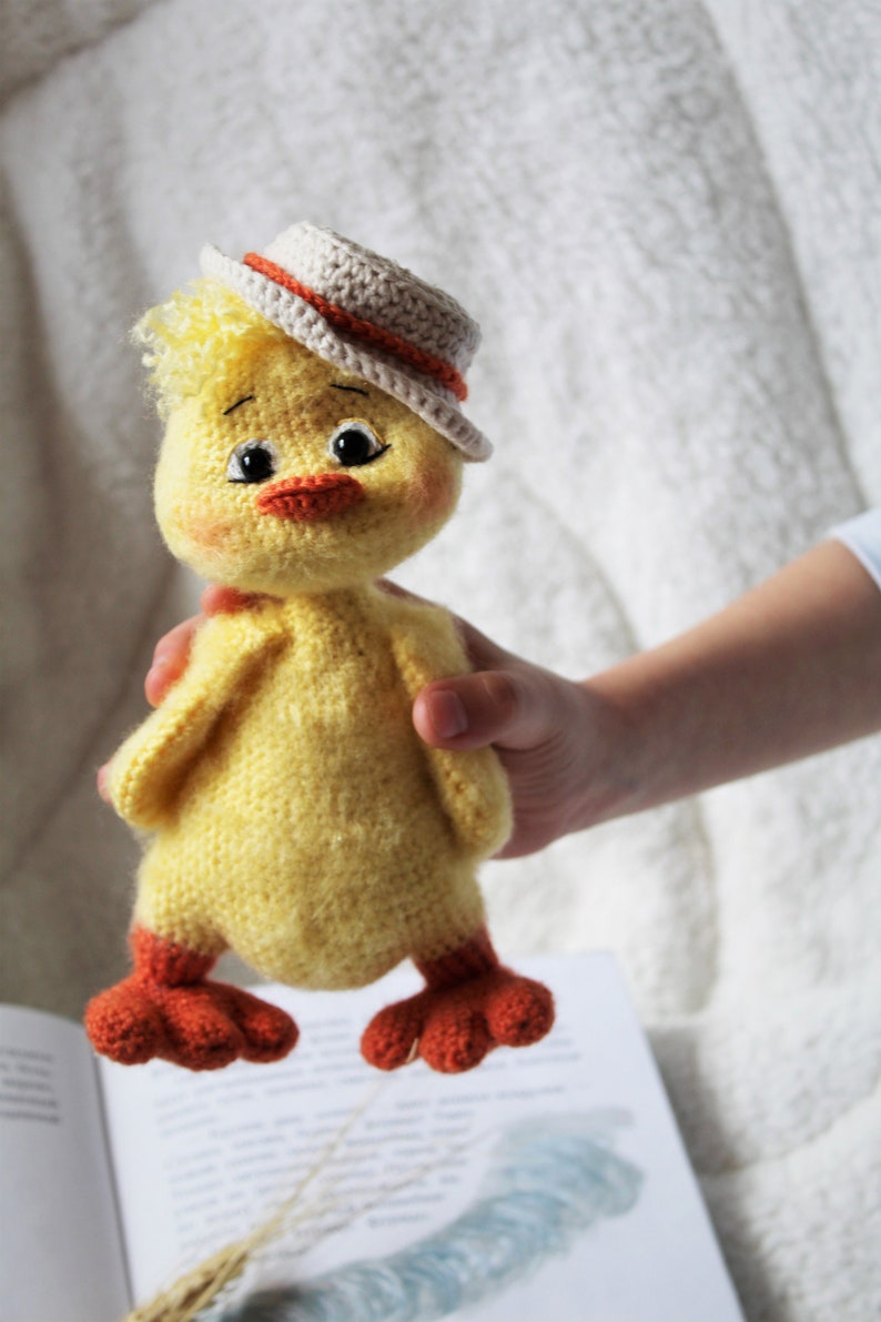 May include: A crocheted yellow chick toy wearing a white and orange hat. The chick has orange feet and black eyes. The chick is being held by a hand.