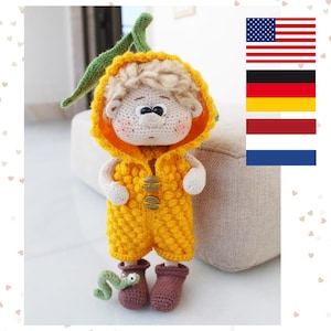 Könnte beinhalten: Eine gehäkelte Puppe, die einen gelben Overall mit einem grünen Wurmspielzeug trägt. Die Puppe hat blonde Haare und eine gelbe Kapuzenjacke.