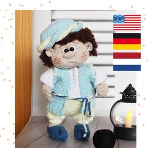 Puede incluir: Una muñeca de crochet con cabello castaño, que lleva un sombrero azul y blanco, un chaleco azul, una camisa blanca, pantalones amarillos y zapatos azules. La muñeca está de pie sobre una superficie de madera con una linterna y lana en el fondo.