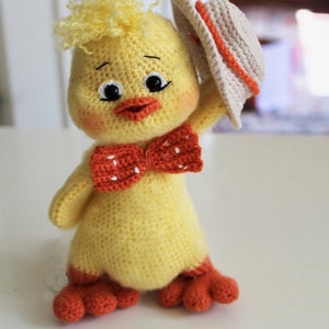 May include: A crocheted yellow chick toy wearing a beige hat and a red bow with white polka dots. The chick has orange feet and a friendly expression.