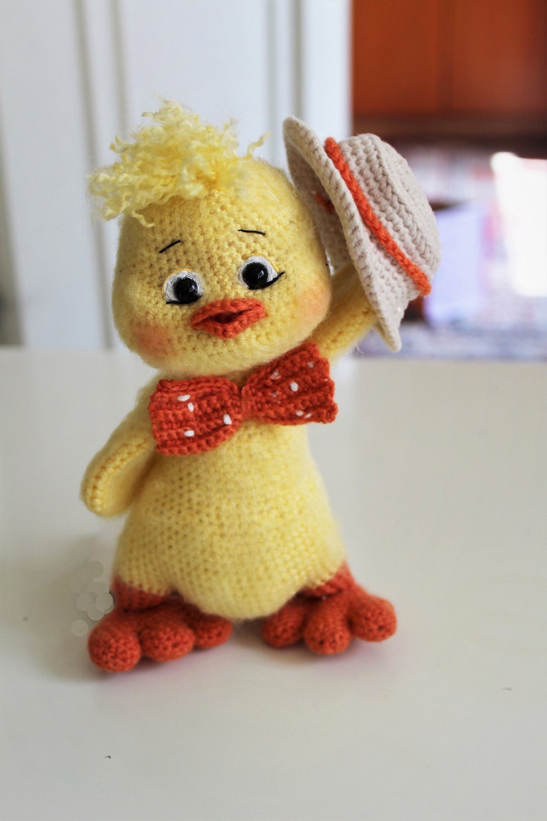 May include: A crocheted yellow chick toy wearing a beige hat and a red bow with white polka dots. The chick has orange feet and a friendly expression.