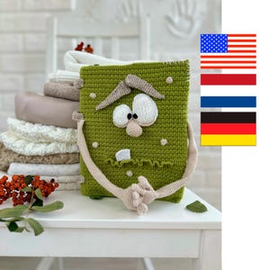 Könnte beinhalten: Eine grüne, gehäkelte Tasche mit Monstergesicht, weißen Augen, einer Nase und einem Zahn. Die Tasche hat beige Arme und Hände und steht auf einem weißen Tisch. Im Hintergrund ein Stapel gefalteter Decken.