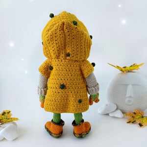 May include: A crocheted doll wearing a yellow hooded jacket with green accents and orange boots. The doll is standing on a white surface with fall leaves and rocks.