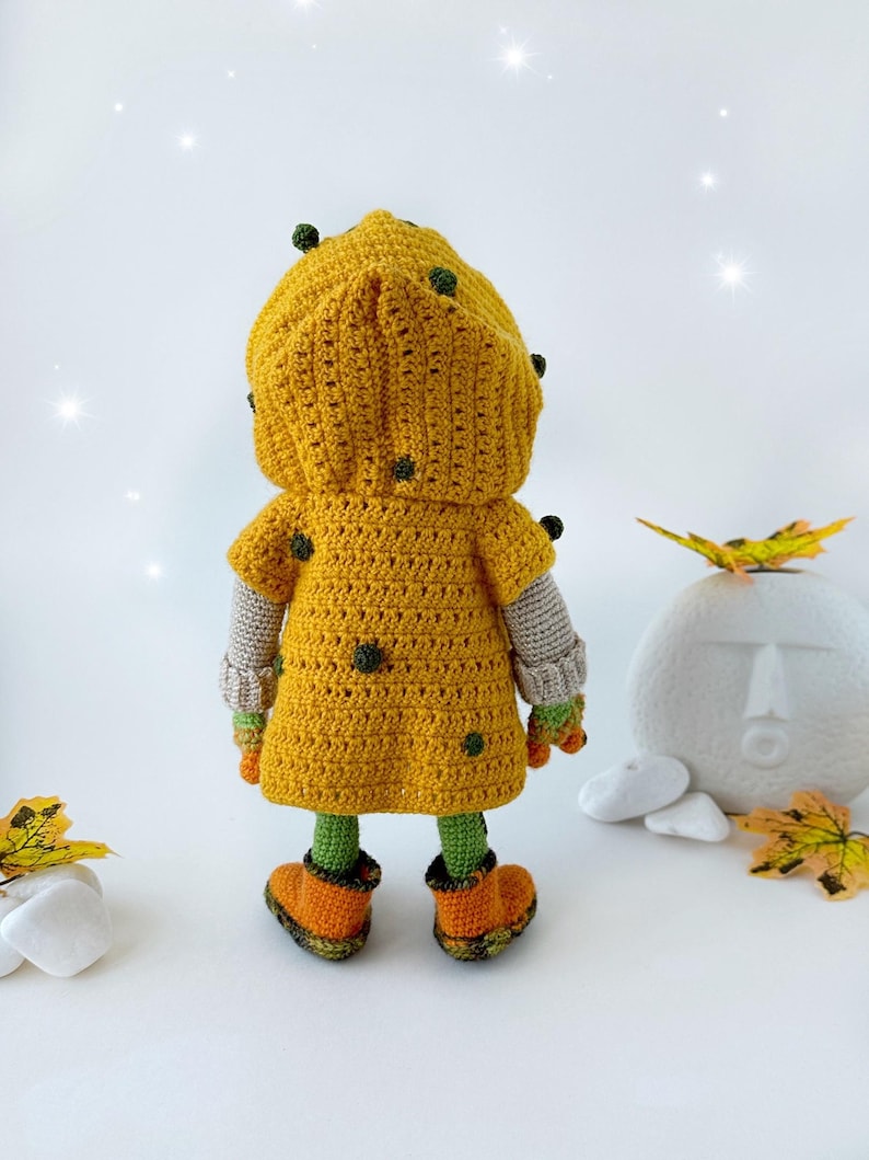 May include: A crocheted doll wearing a yellow hooded jacket with green accents and orange boots. The doll is standing on a white surface with fall leaves and rocks.