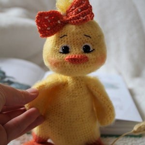 May include: A crocheted yellow chick toy with orange feet and a red and white polka dot bow. The chick has black eyes and an orange beak.