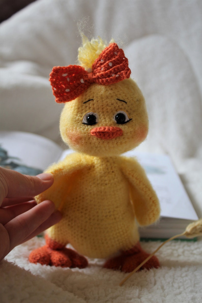 May include: A crocheted yellow chick toy with orange feet and a red and white polka dot bow. The chick has black eyes and an orange beak.