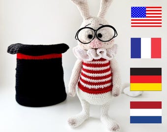 May include: A white crocheted rabbit wearing red and white striped clothing and round glasses. The rabbit is standing next to a black and red top hat.