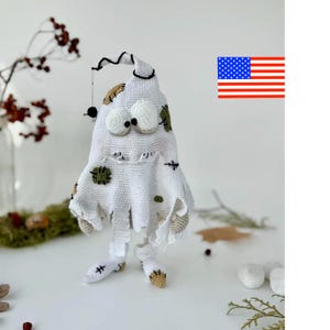 May include: A white crocheted ghost figure with a pointed top, large round eyes, and small arms. The ghost features patches in tan and olive green, black stitched details, and a small black spider hanging from a thread on its head.