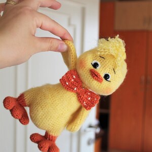 May include: A crocheted yellow chick toy with orange feet and a red and white polka dot bow tie. The chick has a fluffy yellow head and black eyes.