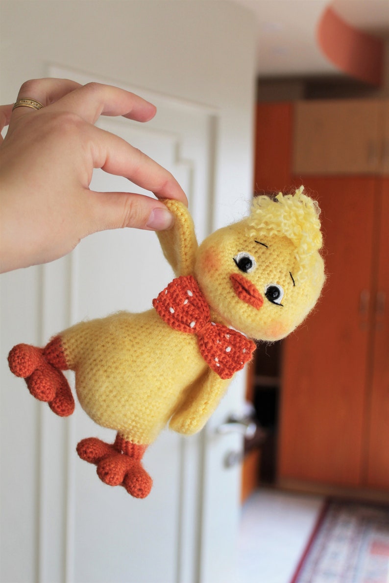 May include: A crocheted yellow chick toy with orange feet and a red and white polka dot bow tie. The chick has a fluffy yellow head and black eyes.