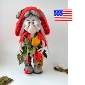 May include: A crocheted doll featuring a large red mushroom cap hood with white spots, long white hair, and a beige face. The doll wears a layered outfit of green, yellow, and red crocheted leaves, with grey boots adorned with small red mushroom accents.
