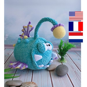 Puede incluir: Un juguete de peluche tejido a crochet con forma de pez pescador azul con un señuelo amarillo. El juguete tiene ojos grandes y blancos y una aleta morada. Está sentado sobre una superficie de madera con una planta verde y piedras.