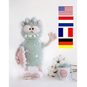 May include: A crocheted green monster toy with pink accents, holding a pink crocheted surfboard. The monster has large eyes, a toothy grin, and long eyelashes. The toy is standing on a white surface with a small white bucket and cotton balls.