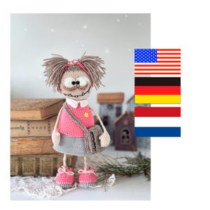 Puede incluir: Muñeca de ganchillo hecha a mano con una parte superior rosa, una falda gris y zapatos a juego. La muñeca tiene un bolso gris, cabello castaño y una expresión caprichosa. La muñeca está cerca de una pila de libros.