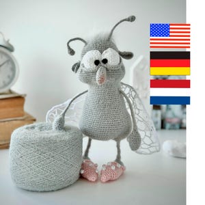 Può includere: Una bambola a forma di insetto grigio all'uncinetto fatta a mano, con grandi occhi bianchi, piedi rosa e ali delicate. Una palla di filo grigio è accanto alla bambola. La bambola ha un design stravagante, perfetto per la cameretta di un bambino.